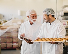 Picture of two employees in sterile white clothes in food factory smiling and talking. Younger man is holding tray full of fresh cookies while the older is holding tablet and checking production line.