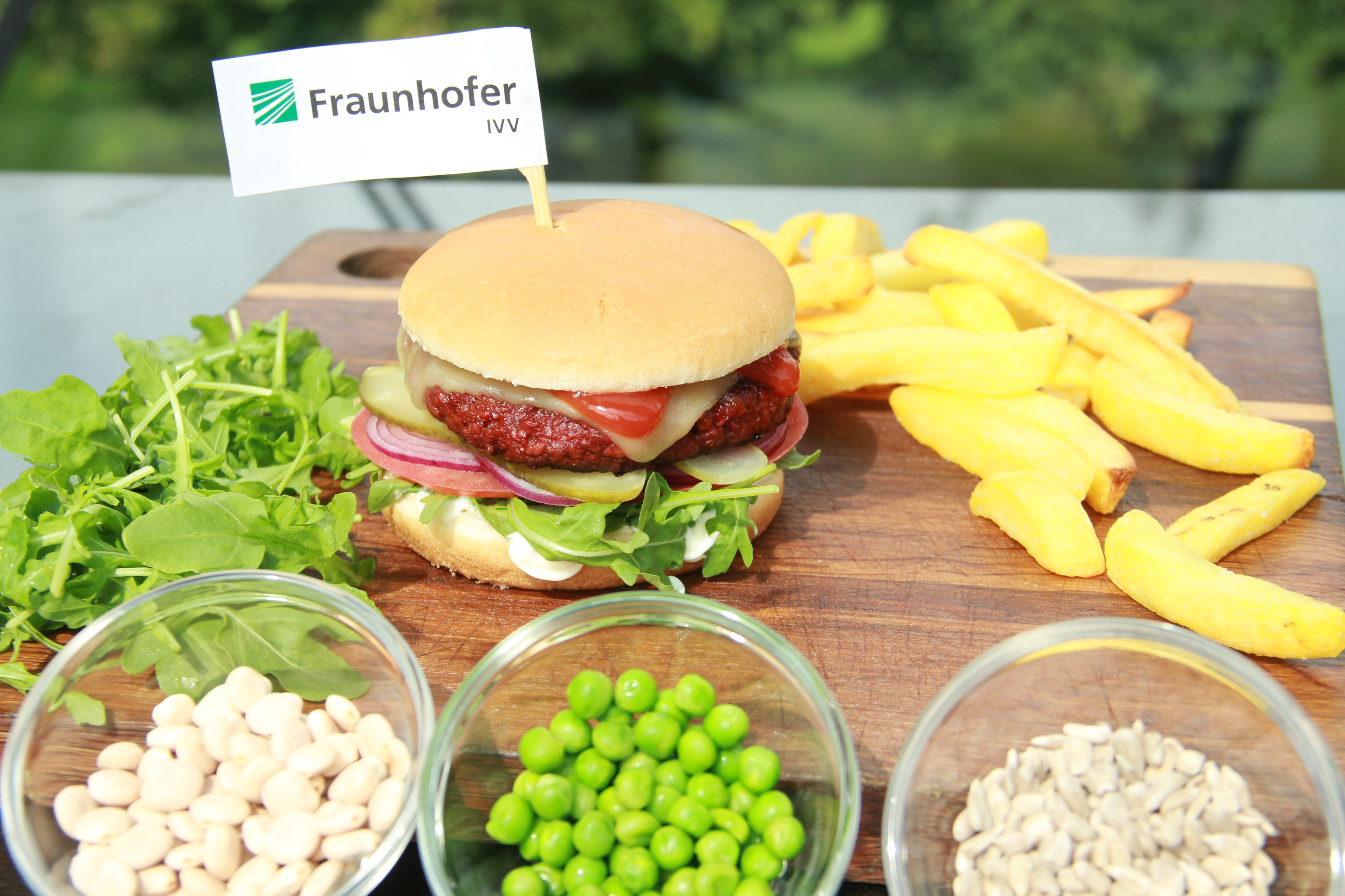 Veganer Burger mit Pommes auf Holzbrett, im Vordergrund drei Schälchen mit den verwendeten Rohstoffen.