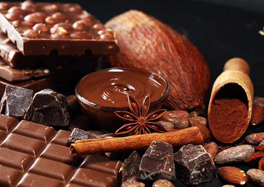 Melted chocolate in a glass dish, cocoa powder in a wooden scoop, a large cocoa bean with shell and small cocoa beans without shell, dark broken chocolate and a stack of chocolate bars decorated with a cinnamon stick and star anise