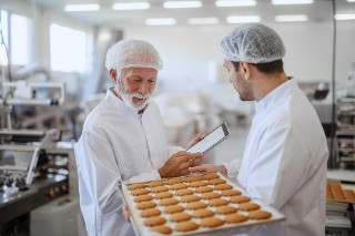 Picture of two employees in sterile white clothes in a food factory, smiling and talking. The younger man is holding a tray of fresh biscuits while the older one is holding a tablet and checking the production line.