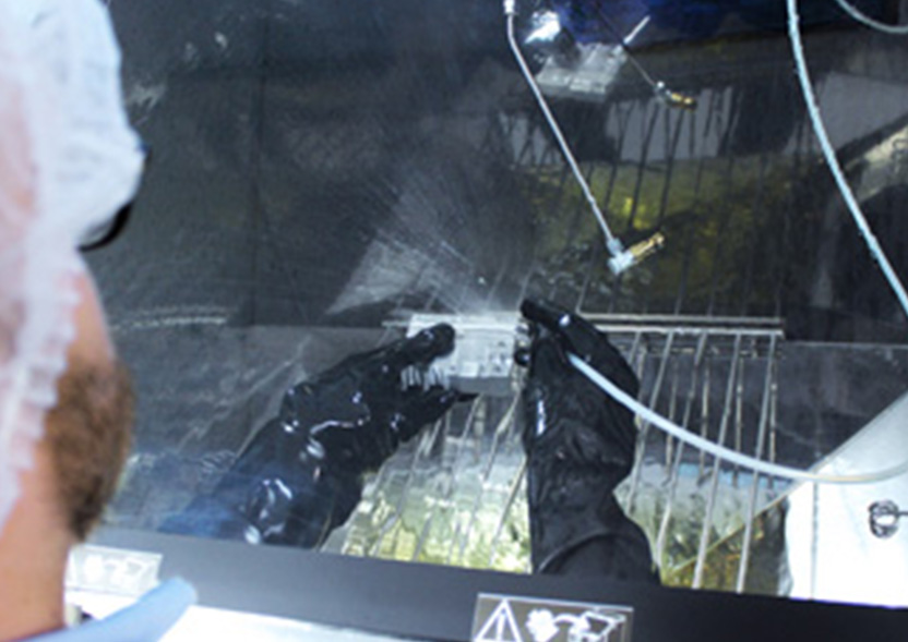 Qualified proof of the technical cleanliness of components according to VDA 19 Part 1 / ISO 16232 A laboratory employee wearing a protective hood and gloves cleans a component in a washing booth. The component and the hands can be seen through a window.