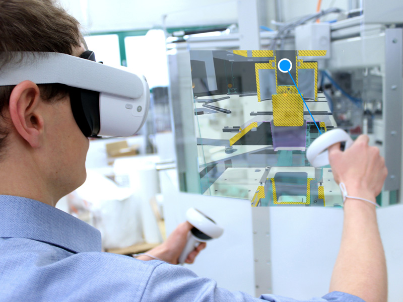 Ortsflexible VR-basierte Qualifikation von Maschinenbedienenden Abbildung eines Mitarbeiters, der mittels VR-Brille virtuell in eine Anlage zur Herstellung tiefgezogener Verpackungen schaut.