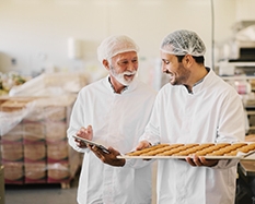 Knowledge management system for artisanal food producers Picture of two employees in sterile white clothes in food factory smiling and talking. Younger man is holding tray full of fresh cookies while the older is holding tablet and checking production line.