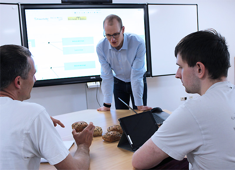 Wissensmanagementsystem für das Lebensmittelhandwerk Bäckereimitarbeitende und ein Forscher des Fraunhofer IVV bei unterhalten sich einem Workshop zur Entwicklung des Wissensmanagementsystems. In der Hand hat ein Mitarbeitender ein Bötchen. Weitere Brötchen liegen auf dem Tisch neben einem Tablet.