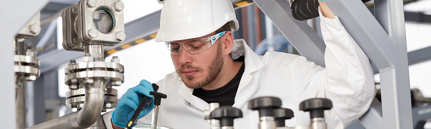 Man in a lab coat, safety goggles and a hard hat in a pilot plant for bio-based products, taking measurements on a machine